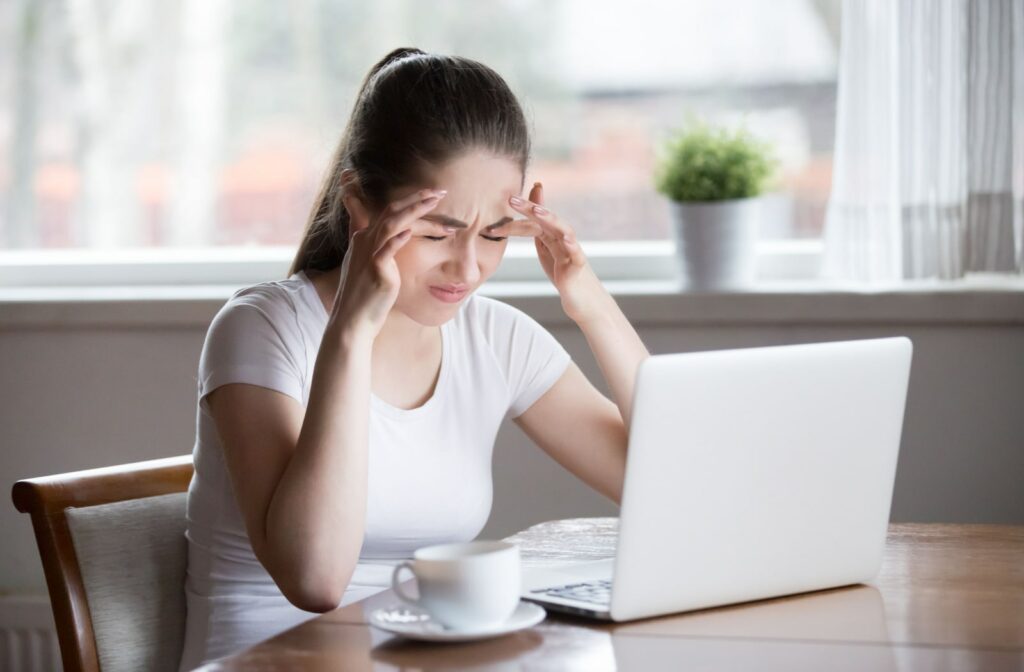 A young woman rubbing her temples and closing her eyes while working in front of an open laptop