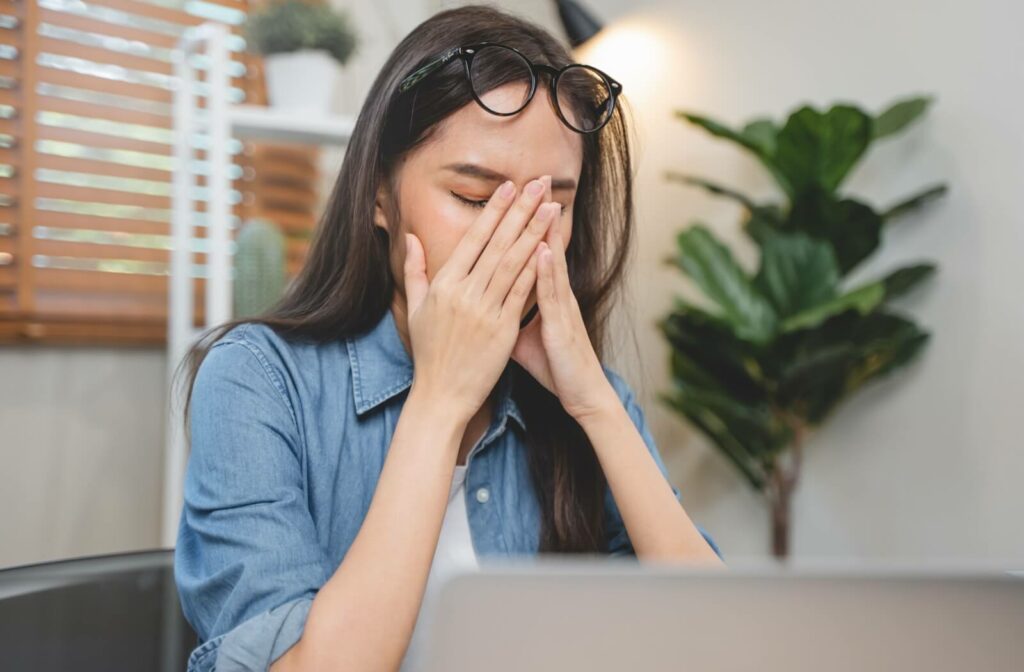 A young woman working at her desk rubbing her eyes with both hands due to dry eye discomfort.