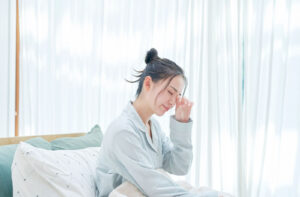 An adult in pyjamas sitting upright in their bed in the morning rubbing their eyes indicating discomfort.