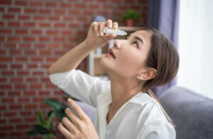 An adult sits on a sofa carefully administering eye drops into their eye.