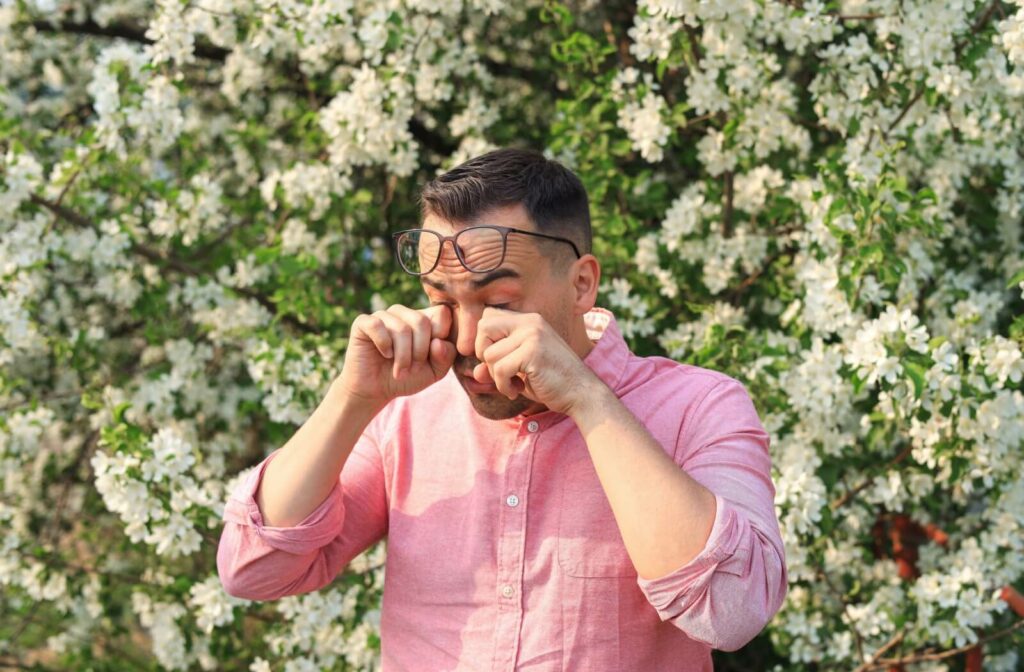 Someone with eye allergies stands in front of a tree and rubs their eyes.