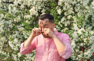 Someone with eye allergies stands in front of a tree and rubs their eyes.