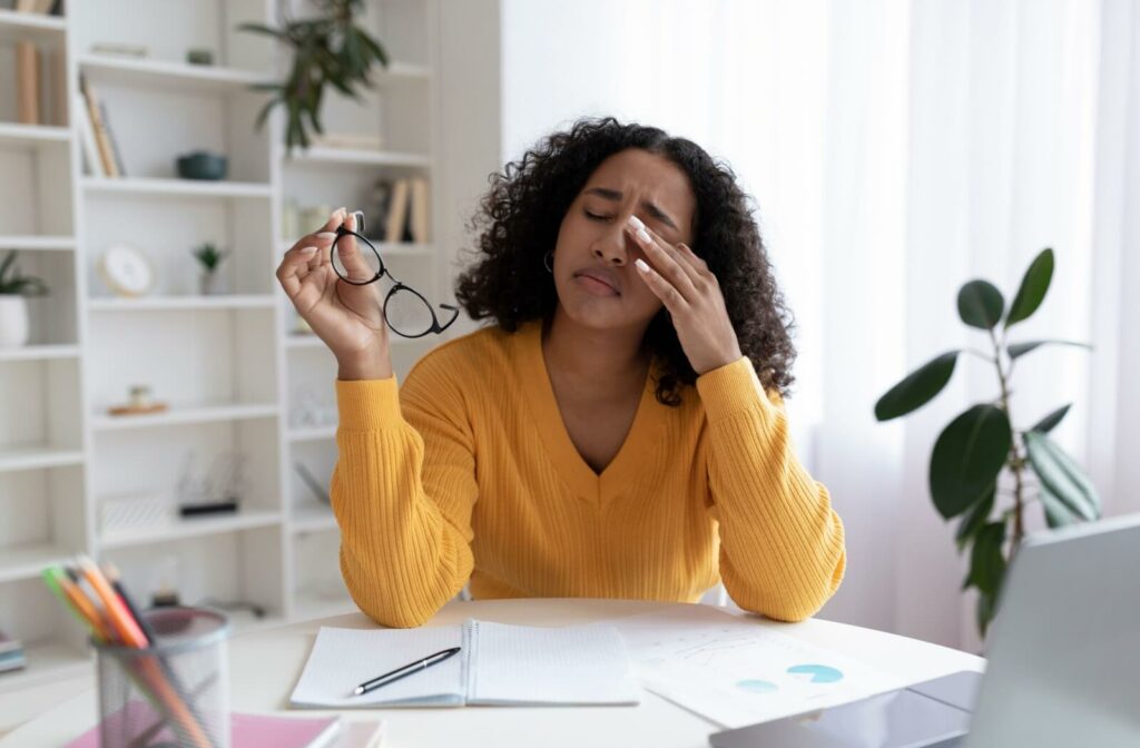 A young woman wearing a yellow sweater sits at a desk, holding her glasses in one hand and gently rubbing her eye with the other, showing signs of eyelid or lash line discomfort.
