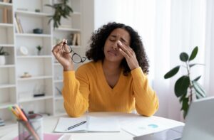 A young woman wearing a yellow sweater sits at a desk, holding her glasses in one hand and gently rubbing her eye with the other, showing signs of eyelid or lash line discomfort.