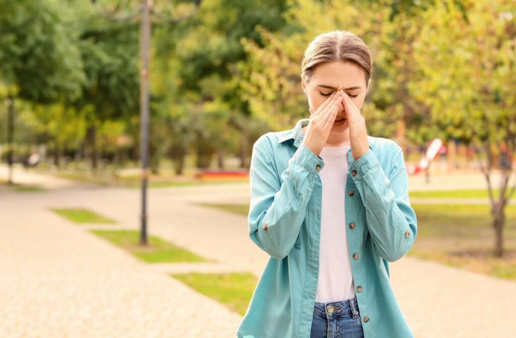 A young woman standing outdoors gently rubbing her eyes with both hands, showing