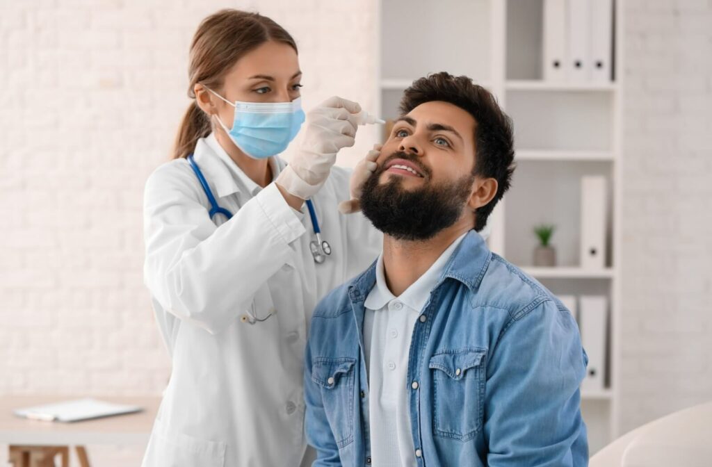 A healthcare provider wearing a mask applies eye drops to a person sitting in a clinic, who is looking upward during the exam.