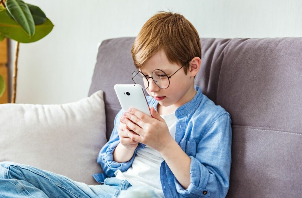 A child with round glasses sits on a couch holding a smartphone close to their face while focusing intently on the screen.