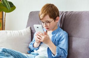 A child with round glasses sits on a couch holding a smartphone close to their face while focusing intently on the screen.