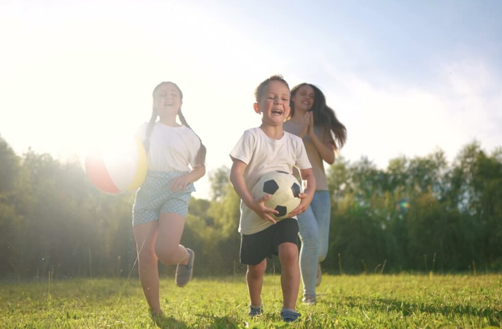 Three children play joyfully outdoors on a sunny day, running through a grassy field with a beach ball and soccer ball.